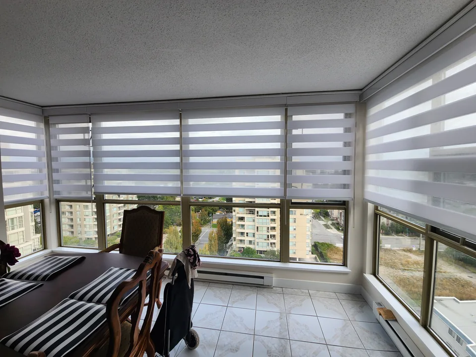 Coordinated white dual shades follow the windows around a dining area, with alternating sheer bands and the lower glazing left visible