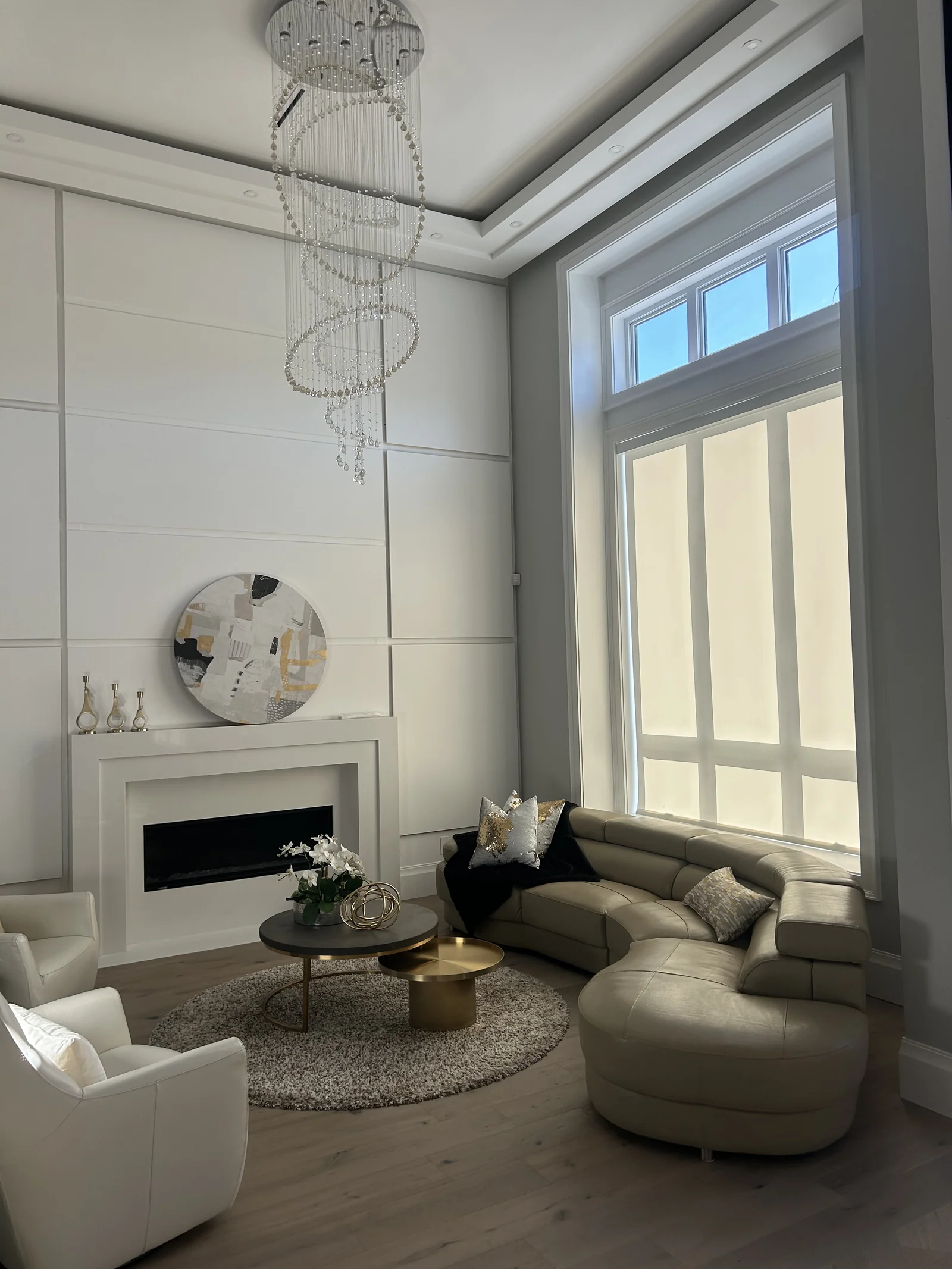 Pale roller shade behind a curved sofa in a tall living room with a fireplace and uncovered upper windows