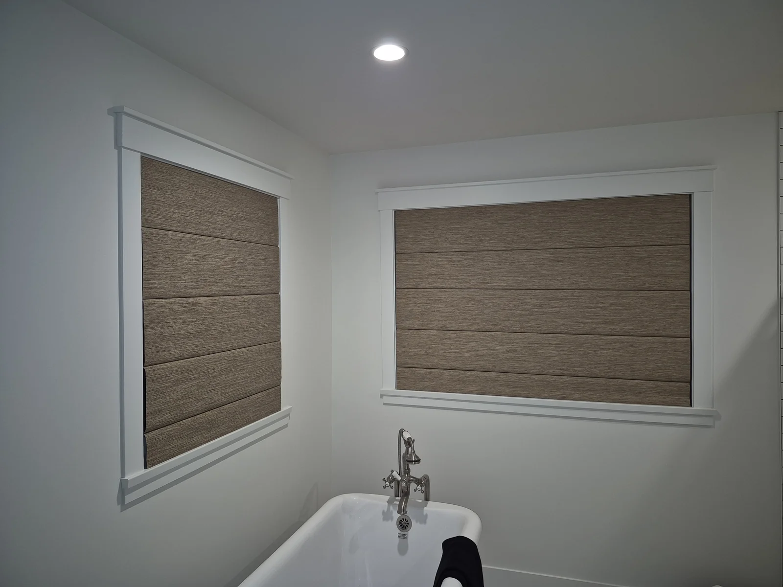 Two warm brown textured Roman shades on adjacent bathroom windows beside a freestanding bath
