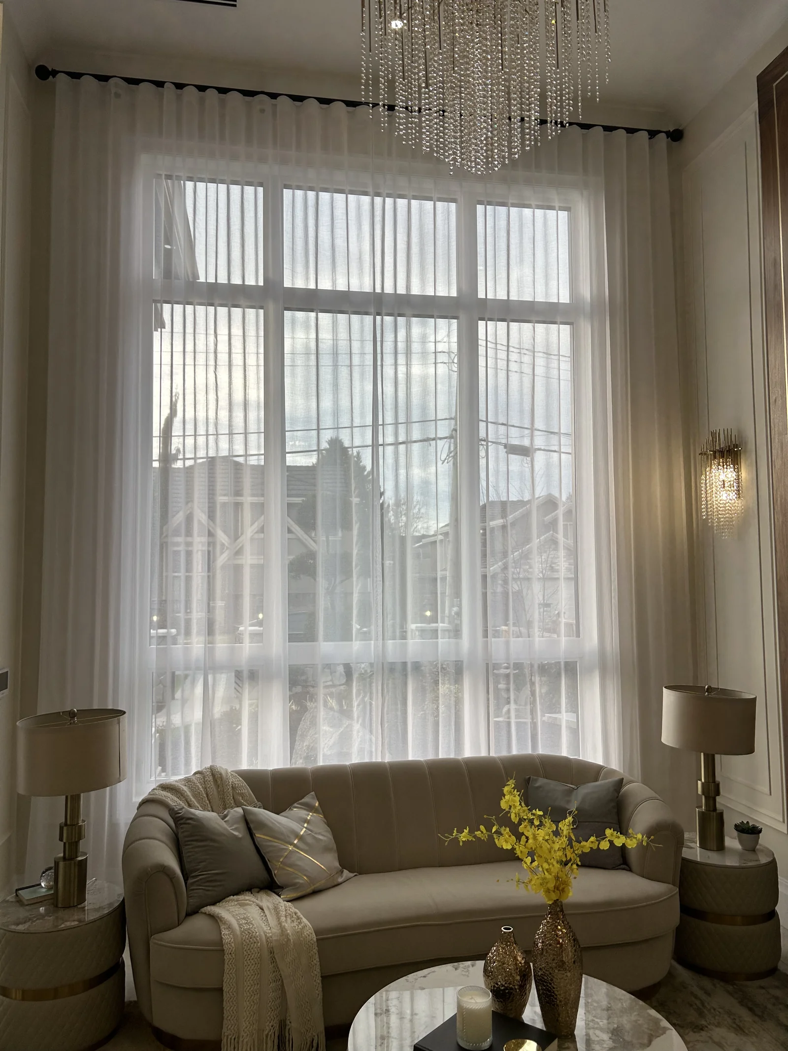 Full-length white sheer curtains at a tall window behind a curved cream sofa and chandelier