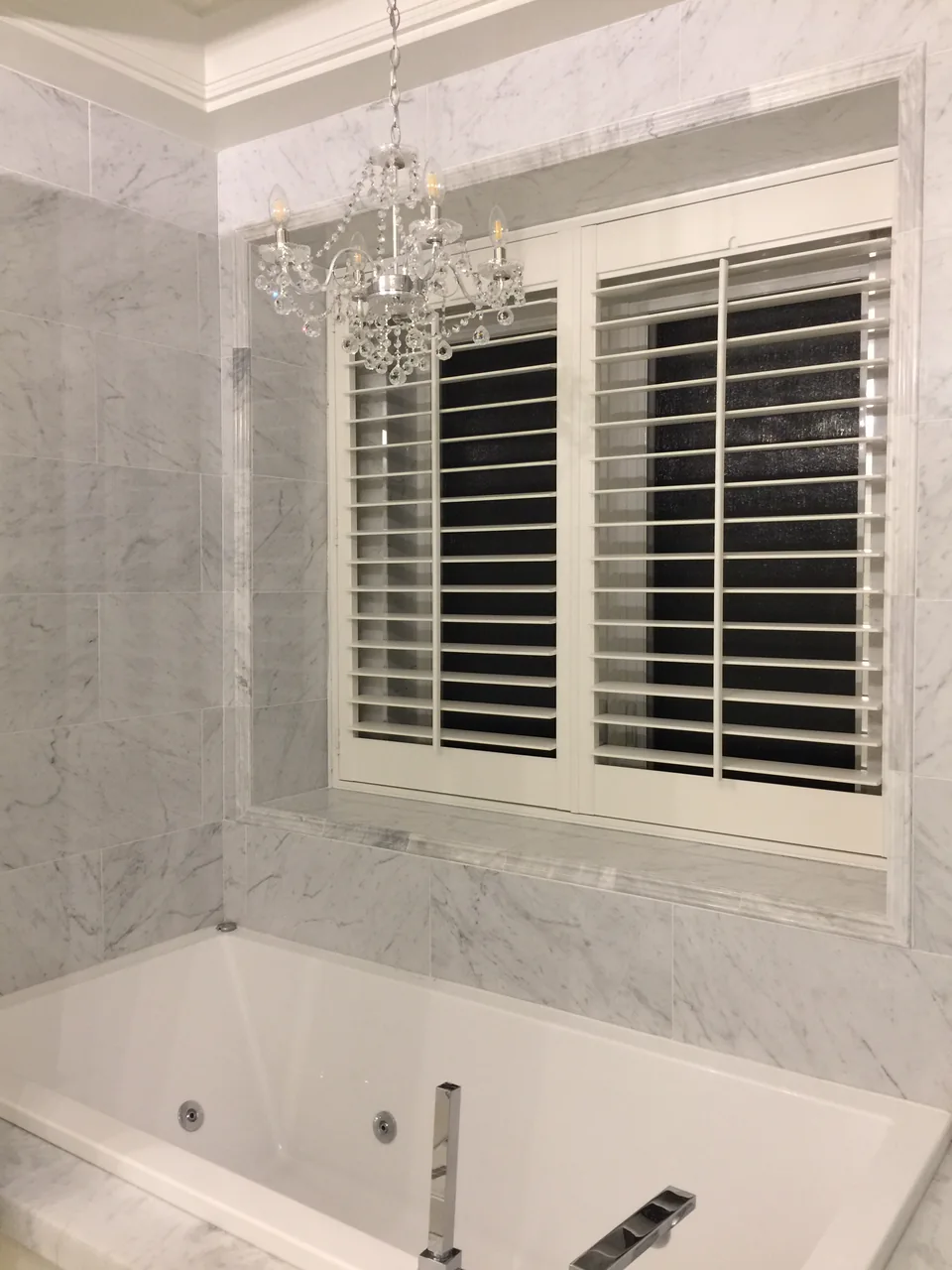 A portrait view of white shutters above a bathtub, framed by marble-patterned walls and a chandelier