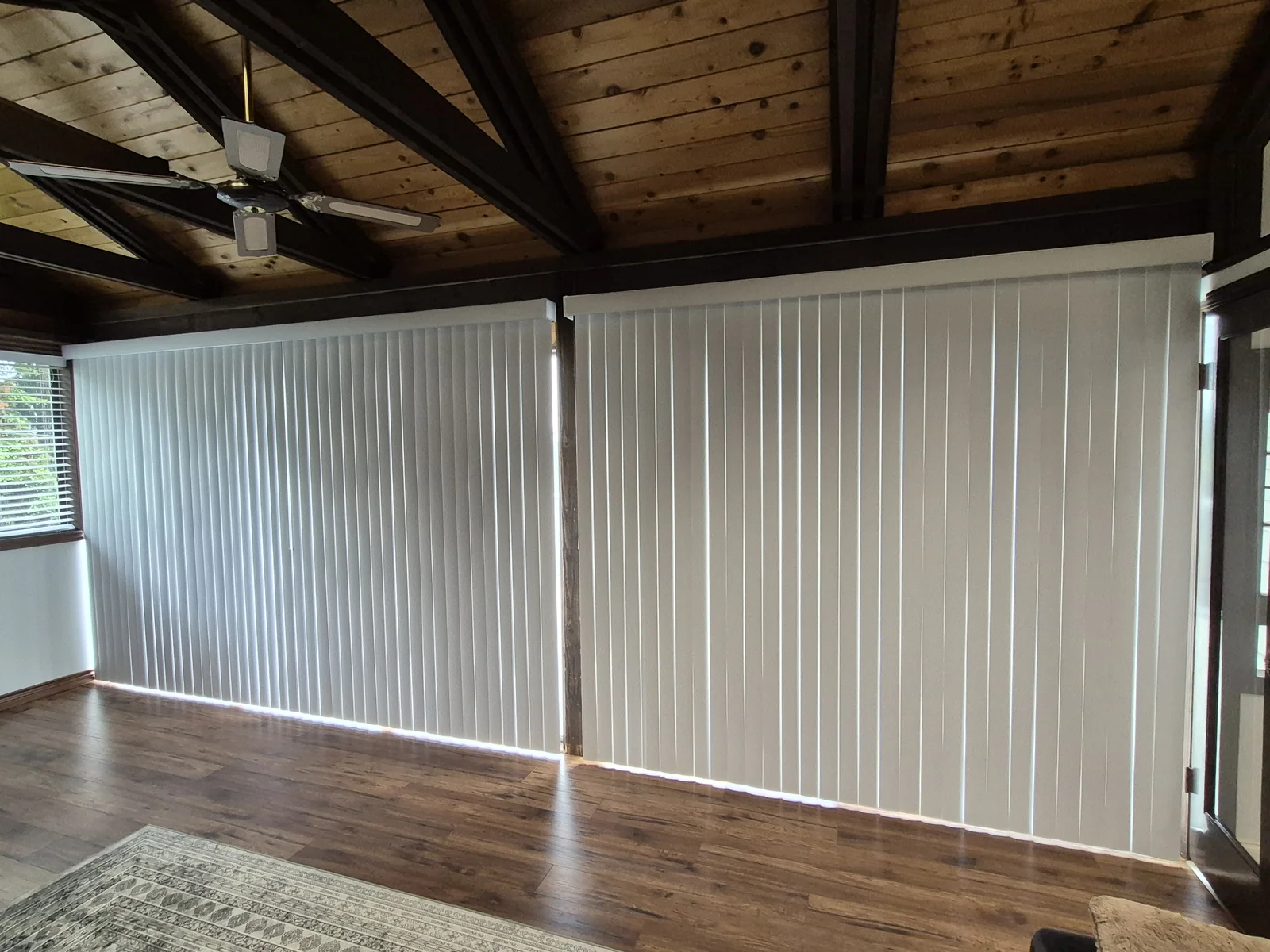 White vertical blinds across wide glazing in a room with a timber ceiling and wood floor