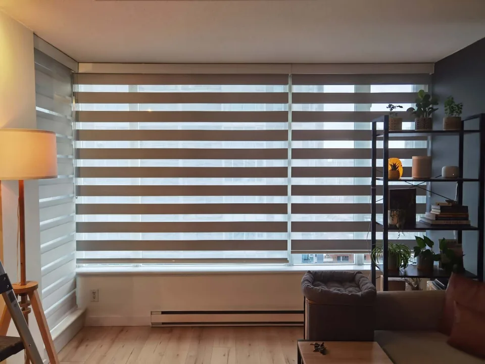 A wide zebra shade brings horizontal bands of filtered light into a living room with wood flooring and warm lamplight